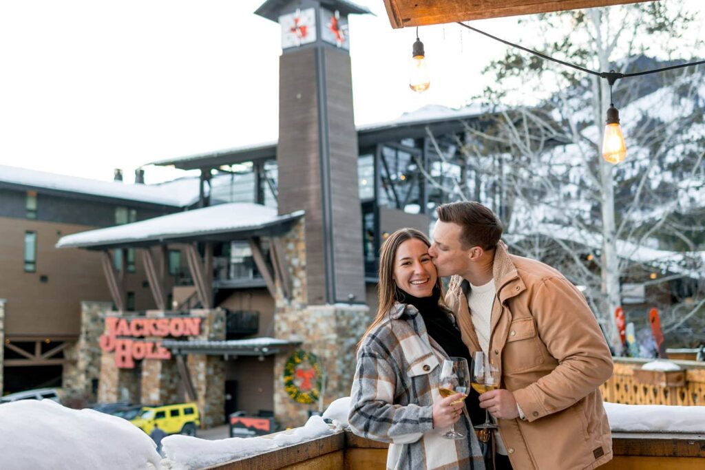 A couple is sharing glasses of wine and a kiss in front of the clock tower at Jackson hole mountain resort