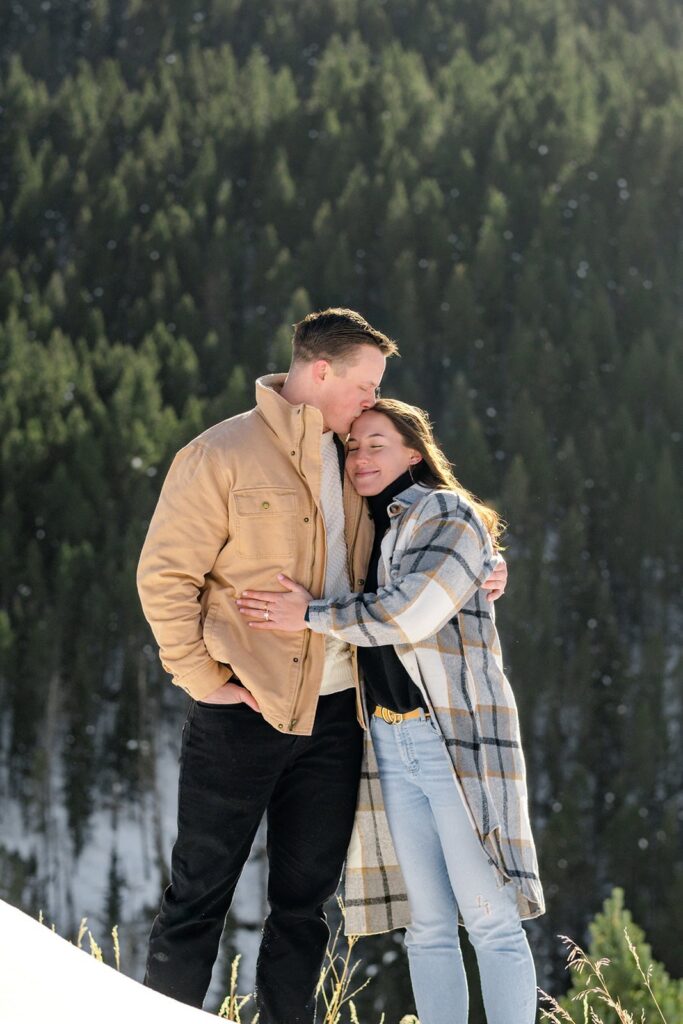 A man kisses his new fiancee's head with pine trees surrounding them