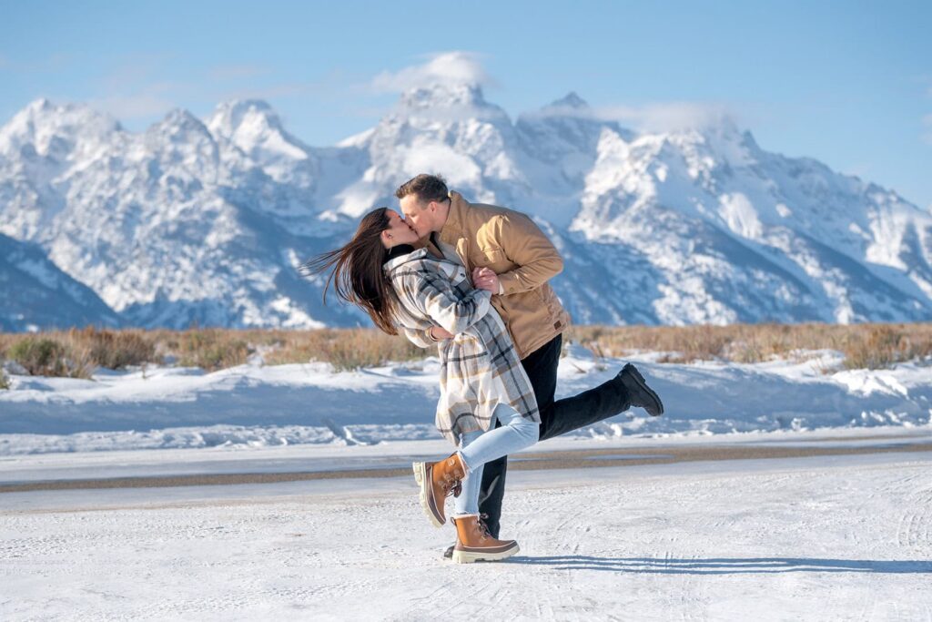 A couple share a dip kiss on a snowy road with the Grand teton mountains behind them. Captured by Jackson hole wedding photographer Karina Dopp Designs